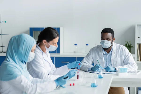 African american scientist with clipboard looking at blurred interracial colleagues with test tubes — Stock Photo