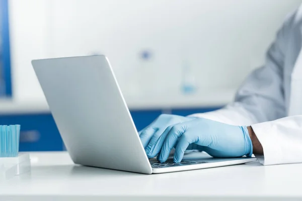 Ausgeschnittene Ansicht eines afrikanisch-amerikanischen Wissenschaftlers in Latexhandschuhen mit Laptop in der Nähe von Reagenzgläsern — Stockfoto