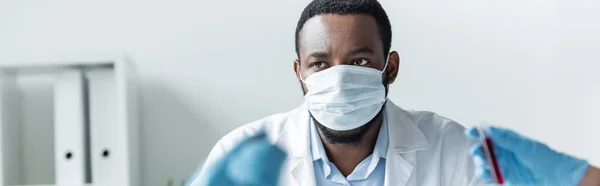 Cientista afro-americano em máscara médica olhando para colegas em primeiro plano borrado, bandeira — Fotografia de Stock