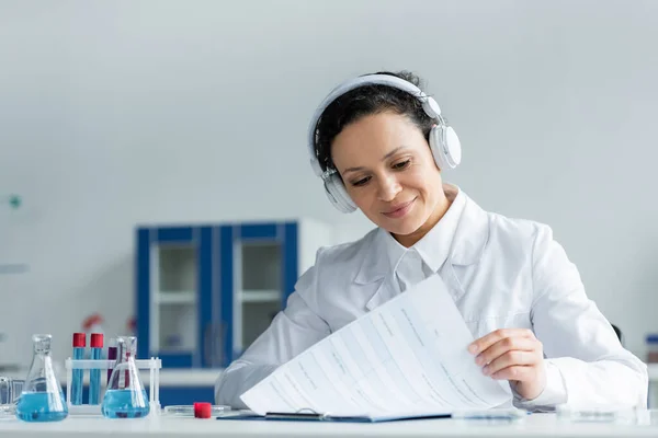 African american scientist in headphones looking at clipboard near test tubes — Stock Photo