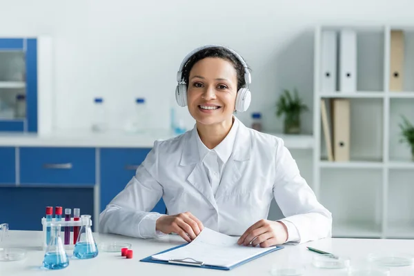 Lächelnder afrikanisch-amerikanischer Wissenschaftler mit Kopfhörern, der in die Kamera neben Klemmbrett und medizinischem Gerät blickt — Stockfoto