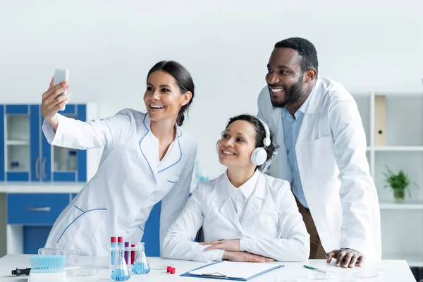 Un scientifique souriant prend du selfie sur smartphone près de collègues multiethniques en laboratoire — Photo de stock