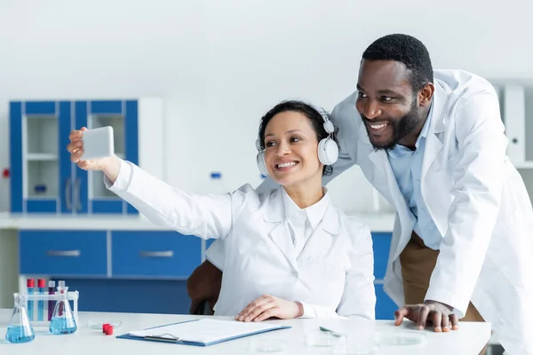 African american scientist in headphones using smartphone near smiling colleague in lab — Stock Photo
