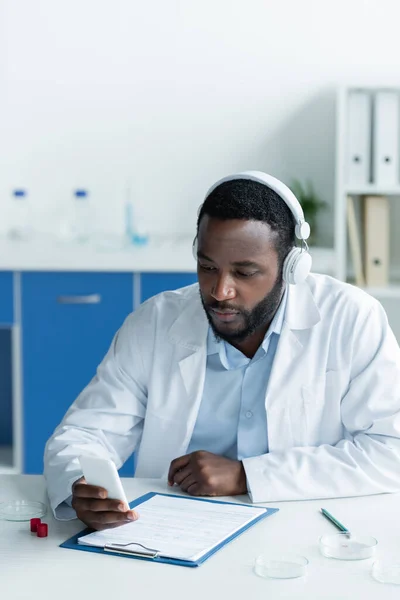 Afrikanischer Wissenschaftler im Kopfhörer mit Smartphone in der Nähe von Petrischalen und Klemmbrett — Stockfoto