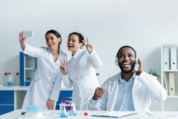 Cientista americano africano alegre em fones de ouvido perto de colegas tirando selfie no laboratório — Fotografia de Stock