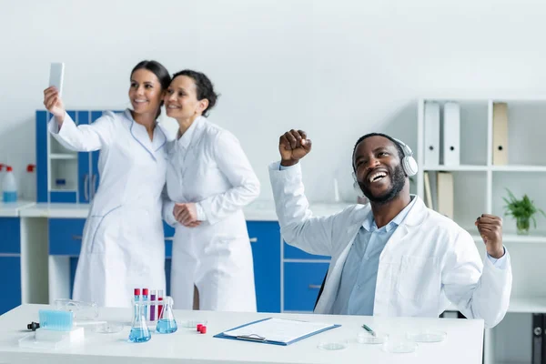Scientifique afro-américain dans les écouteurs montrant un geste oui près de collègues flous avec smartphone dans le laboratoire — Photo de stock