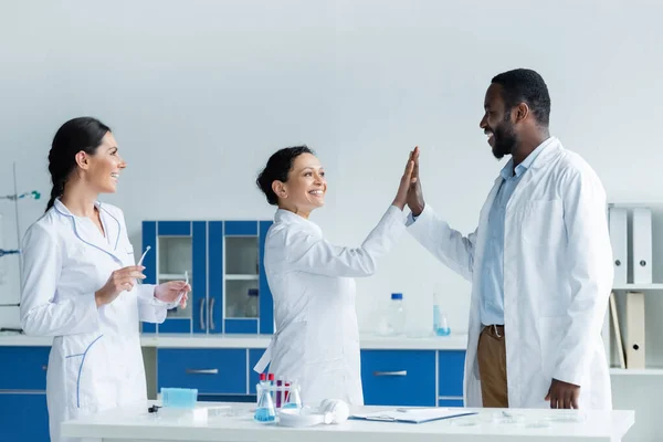 Cientistas afro-americanos sorrindo dando alta cinco perto colega com óculos de segurança no laboratório — Fotografia de Stock