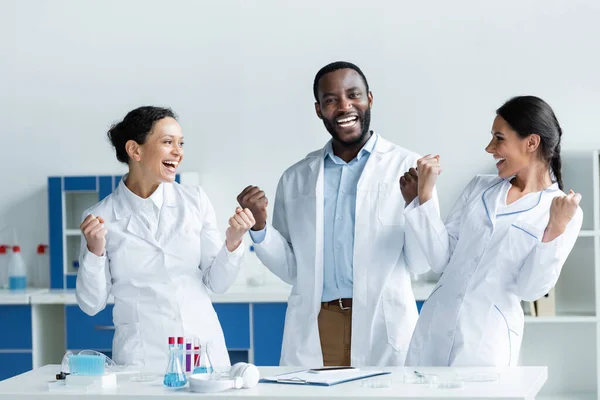 Excited interracial scientists showing yes gesture near smartphone, clipboard and test tubes — Stock Photo