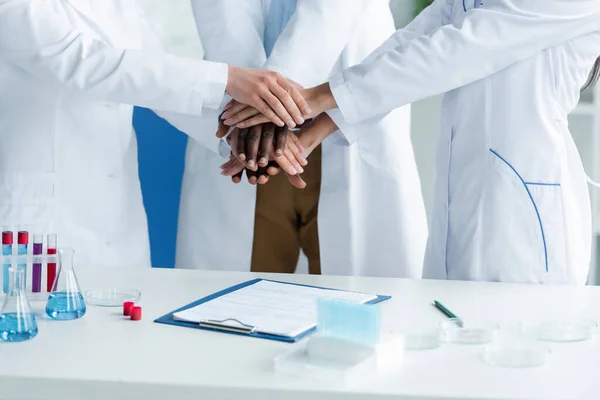 Cropped view of interracial scientists holding hands near clipboard and medical equipment — Stock Photo