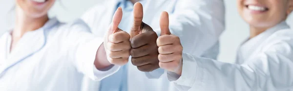 Cropped view of blurred multiethnic doctors showing thumbs up, banner — Stock Photo