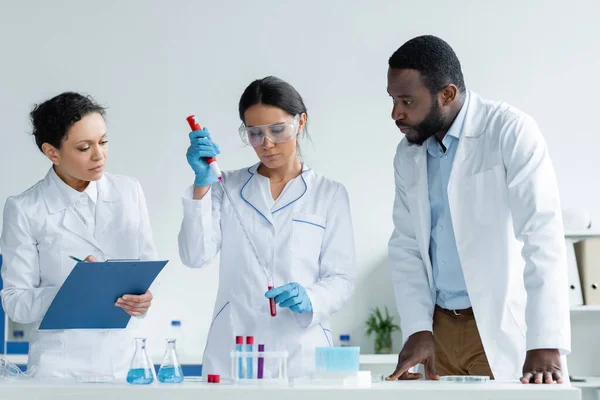 Interracial scientists in white coats working with test tubes and clipboard - foto de stock