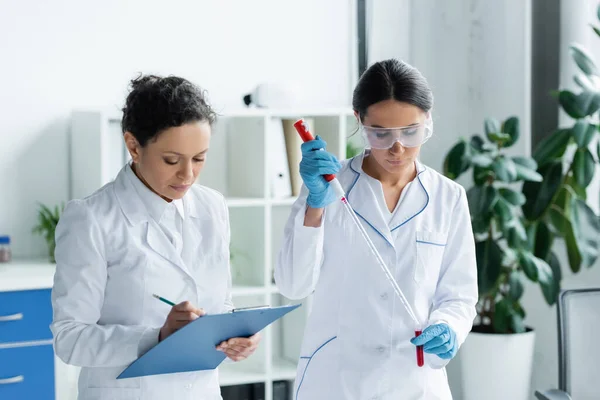 African american scientist writing on clipboard near colleague with test tube and pipette — Stock Photo