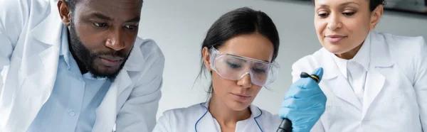 Scientist in goggles holding pipette near interracial colleagues, banner - foto de stock