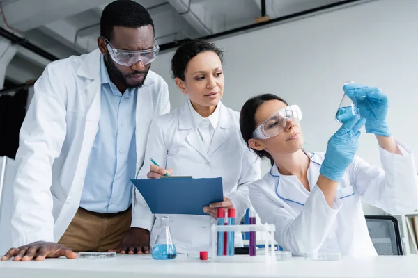 Scientifiques interraciaux en lunettes de sécurité avec presse-papiers regardant le réactif en fiole — Photo de stock