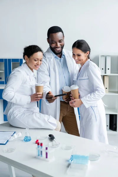 Scienziati interrazziali positivi con caffè guardando smartphone in laboratorio — Foto stock