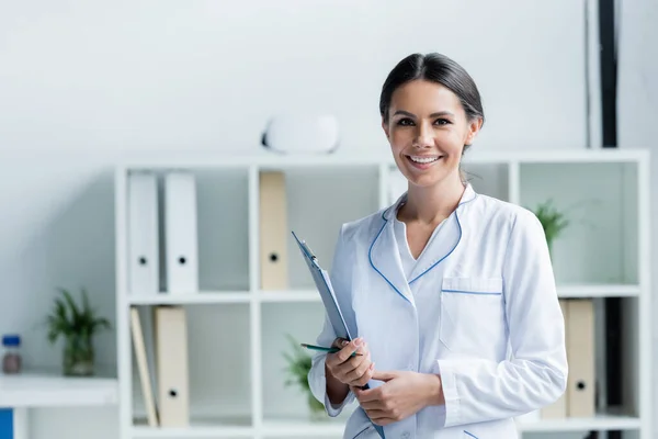 Smiling doctor in white coat holding clipboard in hospital - foto de stock