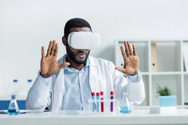 African american scientist in vr headset sitting near blurred medical equipment - foto de stock