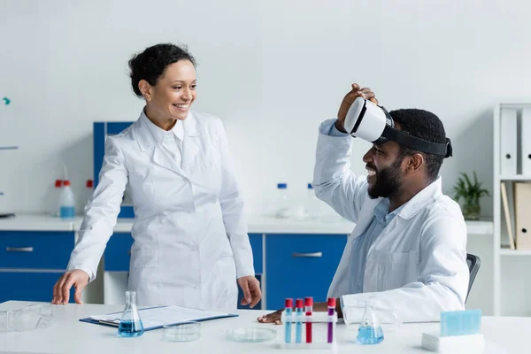 Un scientifique afro-américain debout près d'un collègue dans un casque vr près d'un équipement en laboratoire — Photo de stock