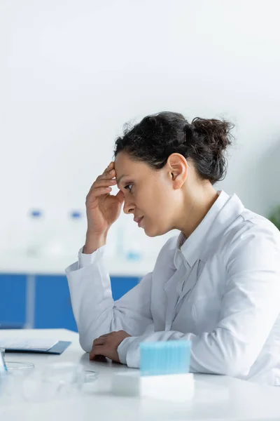 Side view of tired african american scientist sitting near medical equipment — Stock Photo