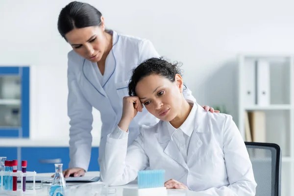 Sad african american scientist sitting near test tubes and colleague on blurred background in lab — Stock Photo