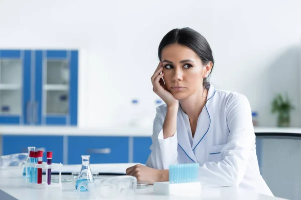Wissenschaftler in weißem Kittel sitzt neben Reagenzgläsern und verschwommener Schutzbrille im Labor — Stockfoto