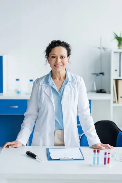 African american scientist smiling at camera near clipboard and medical equipment — Stock Photo