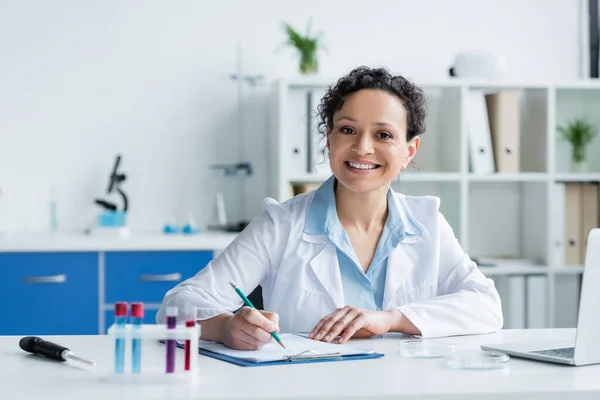 Scienziato afroamericano sorridente che scrive sugli appunti vicino a provette e laptop sfocati — Foto stock