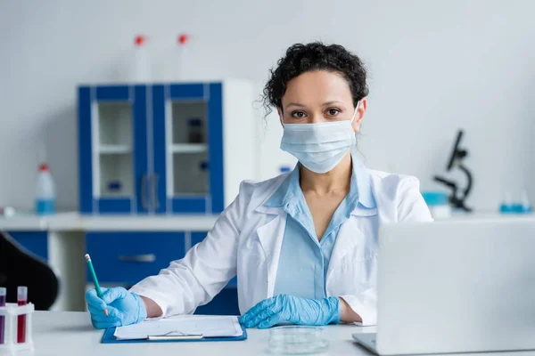 Afrikanischer Arzt in medizinischer Maske und Latexhandschuhen sitzt neben Klemmbrett und Laptop im Labor — Stockfoto