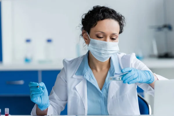 Африканский американский ученый в медицинской маске держит чашку Петри и карандаш возле ноутбука в лаборатории — стоковое фото