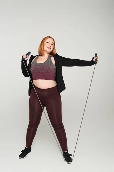 Smiling plus size sportswoman holding corda da salto su sfondo grigio — Foto stock