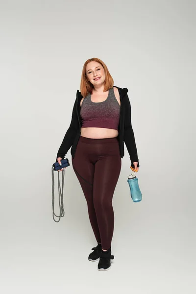 Cheerful plus size sportswoman holding skipping rope and sports bottle on grey background — Stock Photo