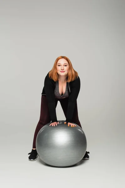 Plus size sportswoman standing near fitness ball on grey background — Stock Photo