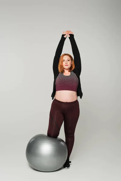 Pelirroja deportista de tamaño grande estirándose cerca de la pelota de fitness sobre fondo gris - foto de stock