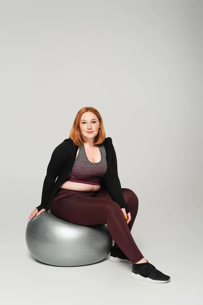 Deportiva de talla grande sentada en una pelota de fitness y mirando a la cámara sobre fondo gris - foto de stock