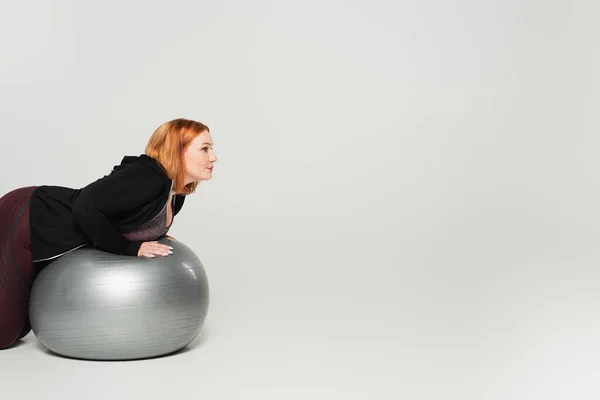 Боковой вид плюс размер женщины, тренирующейся на фитнес-мяче на сером фоне — стоковое фото