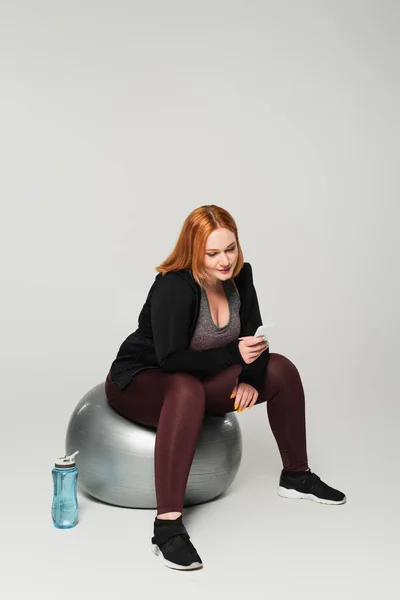 Corpo sportivo positivo utilizzando smartphone sulla palla fitness vicino a bottiglia sportiva su sfondo grigio — Foto stock