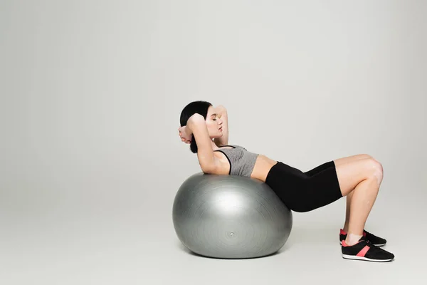 Vista lateral de la deportista con vitiligo haciendo ejercicio sobre pelota de fitness sobre fondo gris - foto de stock