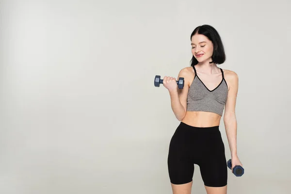 Cheerful sportswoman with vitiligo working out with dumbbells isolated on grey — Stock Photo