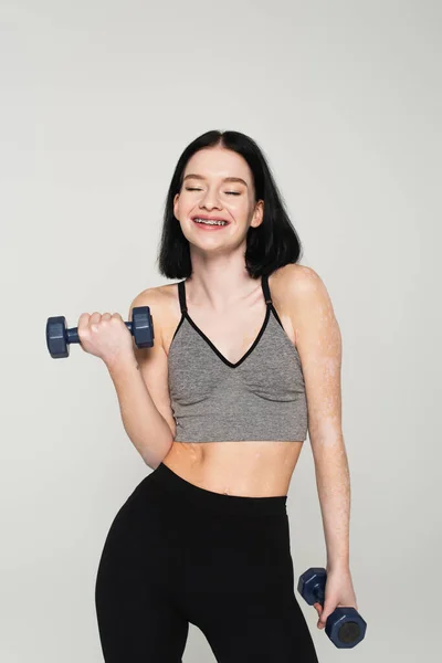 Positive sportswoman with vitiligo working out with dumbbells isolated on grey — Stock Photo