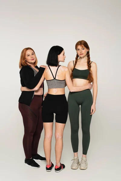 Körperpositive Frauen in Sportbekleidung umarmen sich auf grauem Hintergrund — Stockfoto