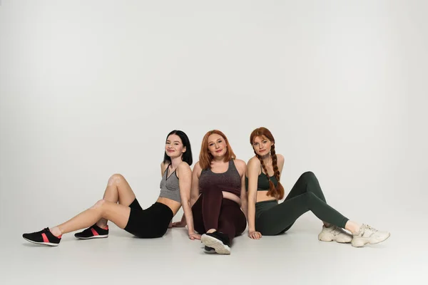 Smiling body positive women in sportswear looking at camera on grey background — Stock Photo