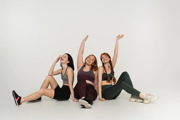 Cheerful body positive sportswomen sitting on grey background — Stock Photo