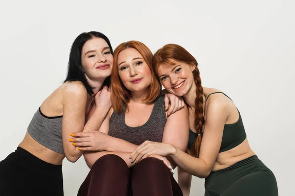 Cheerful body positive sportswomen hugging plus size friend isolated on grey — Stock Photo