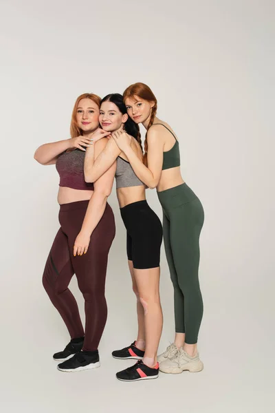 Longitud completa de mujeres sonrientes cuerpo positivo en ropa deportiva abrazándose unos a otros sobre fondo gris - foto de stock