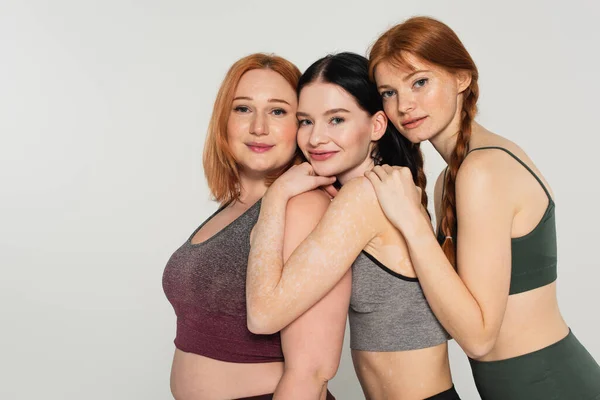 Young body positive friends in sportswear hugging isolated on grey — Stock Photo