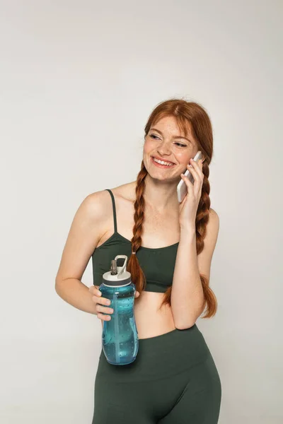 Усміхнена веснянка спортсменка говорить на смартфоні і тримає спортивну пляшку ізольовано на сірому — стокове фото