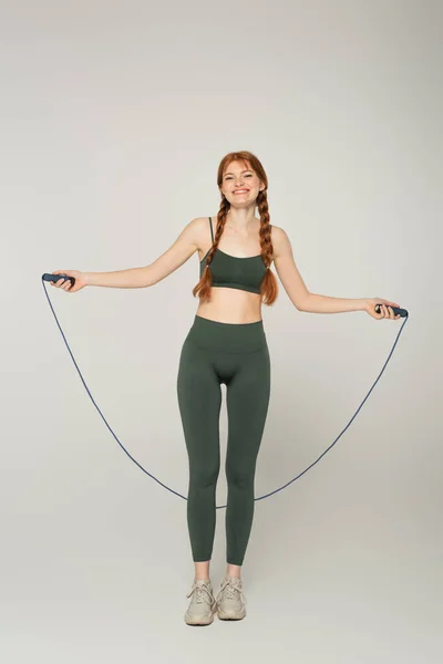 Deportista sonriente y pecosa sosteniendo cuerda de salto sobre fondo gris - foto de stock