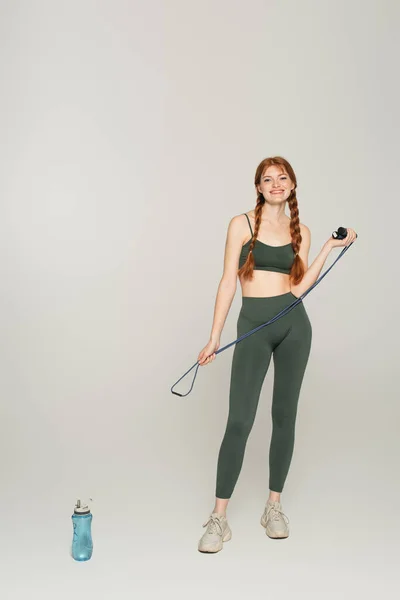 Femme sportive joyeuse avec des taches de rousseur tenant corde à sauter près de la bouteille de sport sur fond gris — Photo de stock