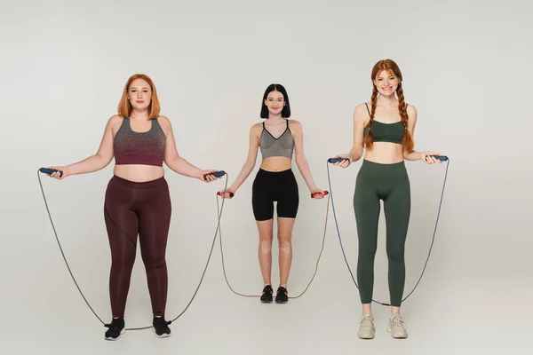 Smiling body positive sportswomen holding jump ropes on grey background — Stock Photo