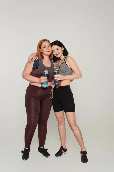 Sportswoman con vitiligine abbracciare amico con bottiglia sportiva e saltare corda su sfondo grigio — Foto stock